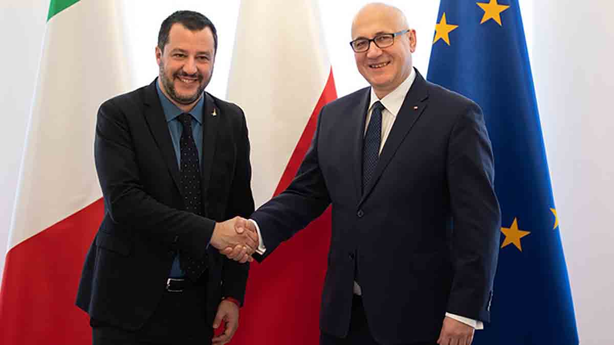 Wicepremier Włoch Matteo Salvini i szef MSWiA Joachim Brudziński podczas spotkania w Warszawie ściskają sobie dłonie na tle flag Włoch, Polski i Unii Europejskiej