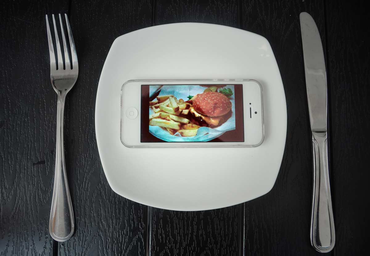 Telefon komórkowy na talerzu  z wyświetlonym hamburgerem. Obok talerza leżą sztućce 