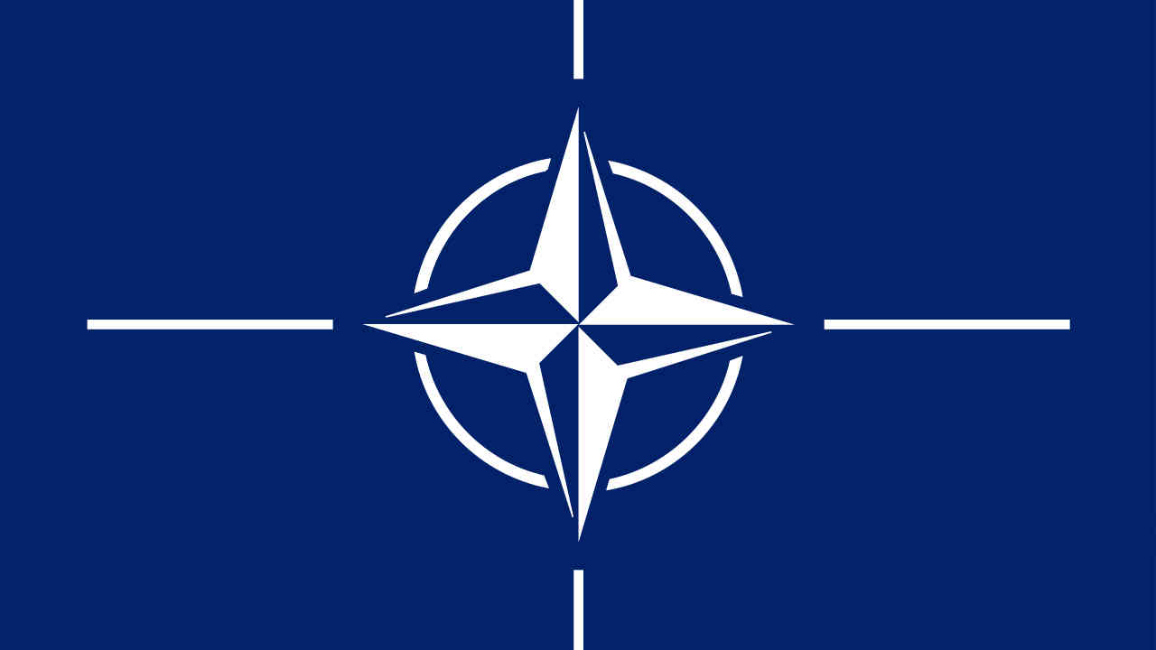 Flaga NATO czteroramienna gwiazda w kole, od ramion w poziomie i pionie odchodzą przedłużenia linii po odstępie. Całość przypomina celownik snajperski