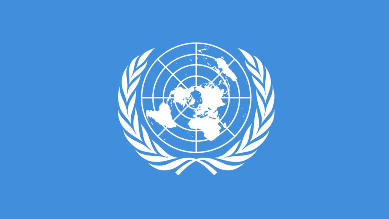 Flaga ONZ, widok planu Ziemi od strony bieguna północnego rzuconego na siatkę w kole, Ziemia na siatce otoczona wieńcem laurowym z przerwą na górze. Wszystko na jasnoniebieskim tle.